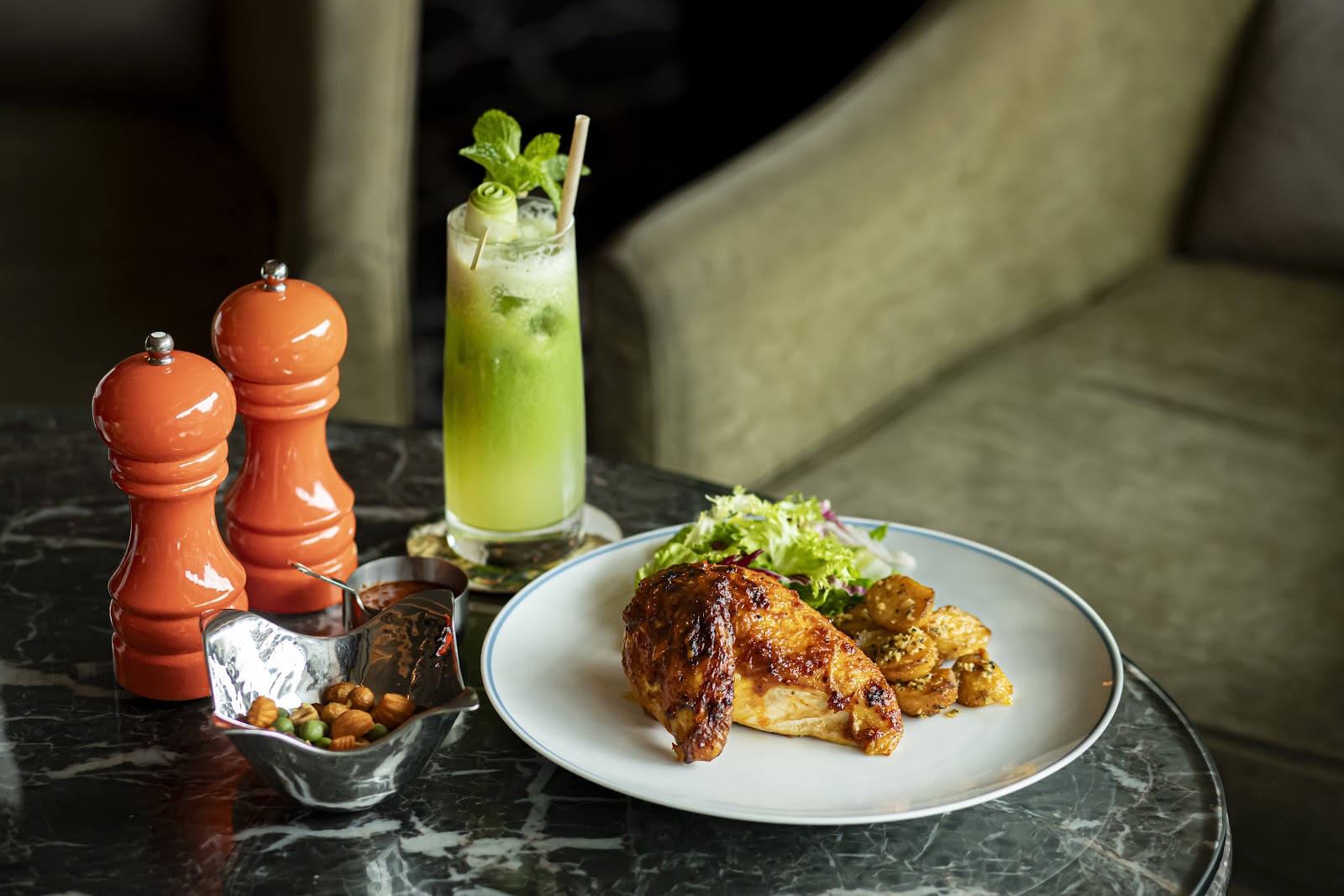 Đĩa gà quay da giòn kèm khoai và salad trên bàn đá, ly nước xanh bạc hà và đôi lọ xay tiêu cam