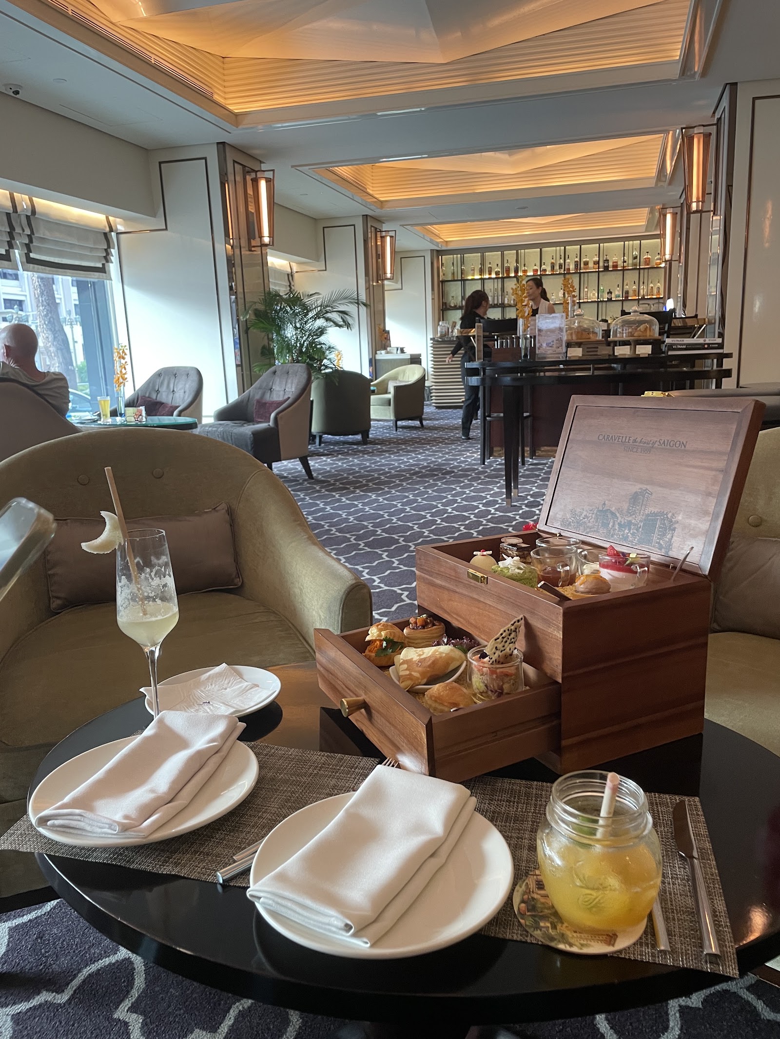 Trà chiều trong hộp gỗ khắc 'Caravelle — the heart of Saigon': bánh mặn, bánh ngọt và ly tráng miệng, nhìn ra quầy bar