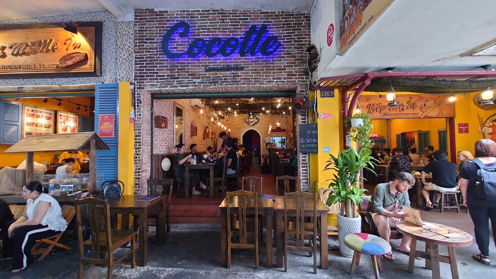 Quán lúc ban ngày: bàn gỗ ngoài vỉa hè, thực khách đông, biển neon Cocotte phía trên