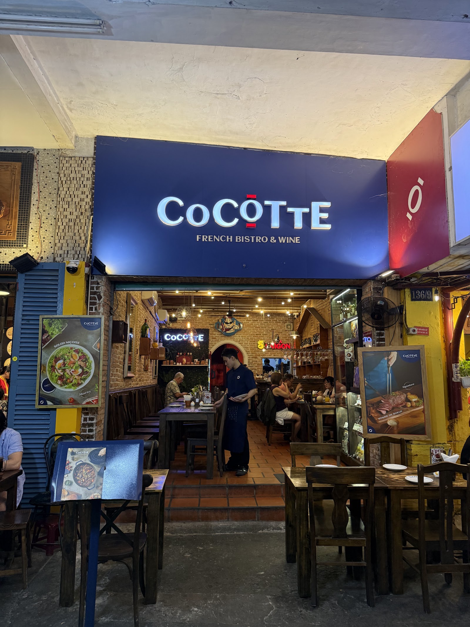 Biển hiệu xanh 'COCOTTE FRENCH BISTRO & WINE' trên cửa ra vào, hai bảng áp phích quảng cáo salade niçoise và rosbif de boeuf bên cạnh
