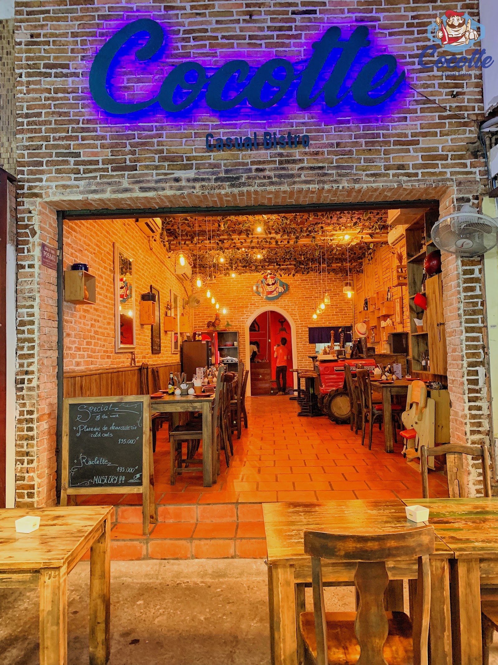 Mặt tiền quán lúc lên đèn: tường gạch, biển neon xanh chữ 'Cocotte Casual Bistro', bảng phấn 'món đặc biệt của tuần' ngay cửa và bàn gỗ ngoài vỉa hè