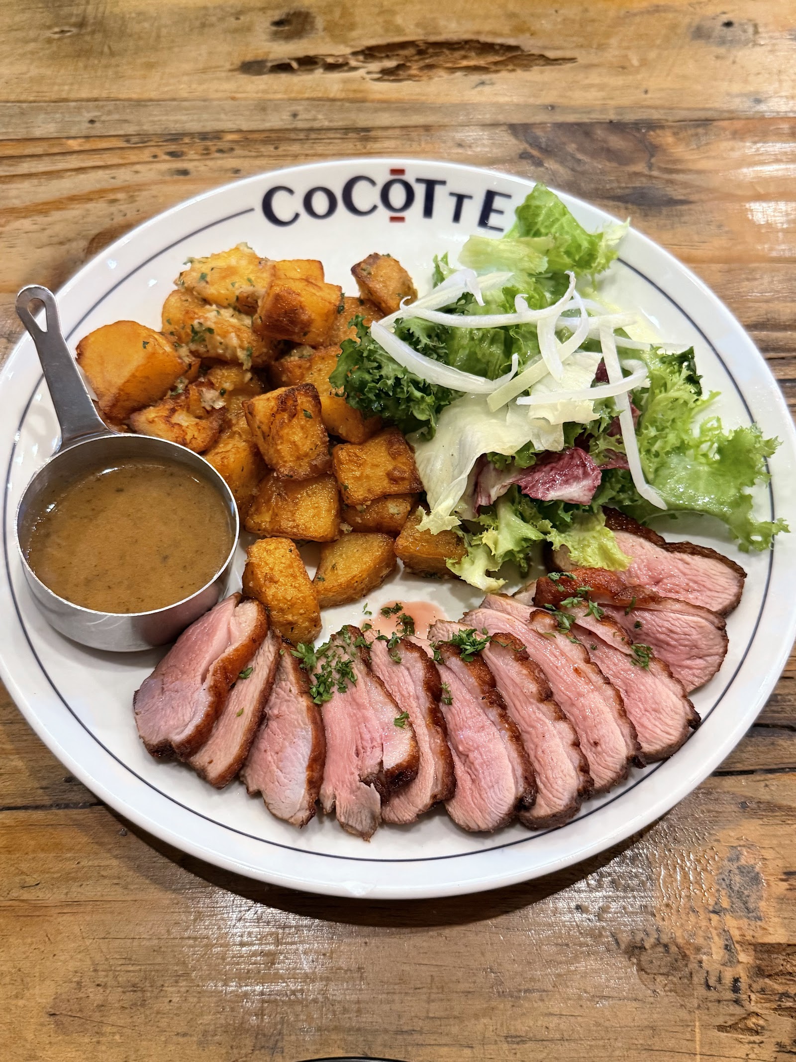 Đĩa vịt thái lát da giòn, khoai tây nướng, salad xanh và chén sốt tiêu trên đĩa in tên Cocotte