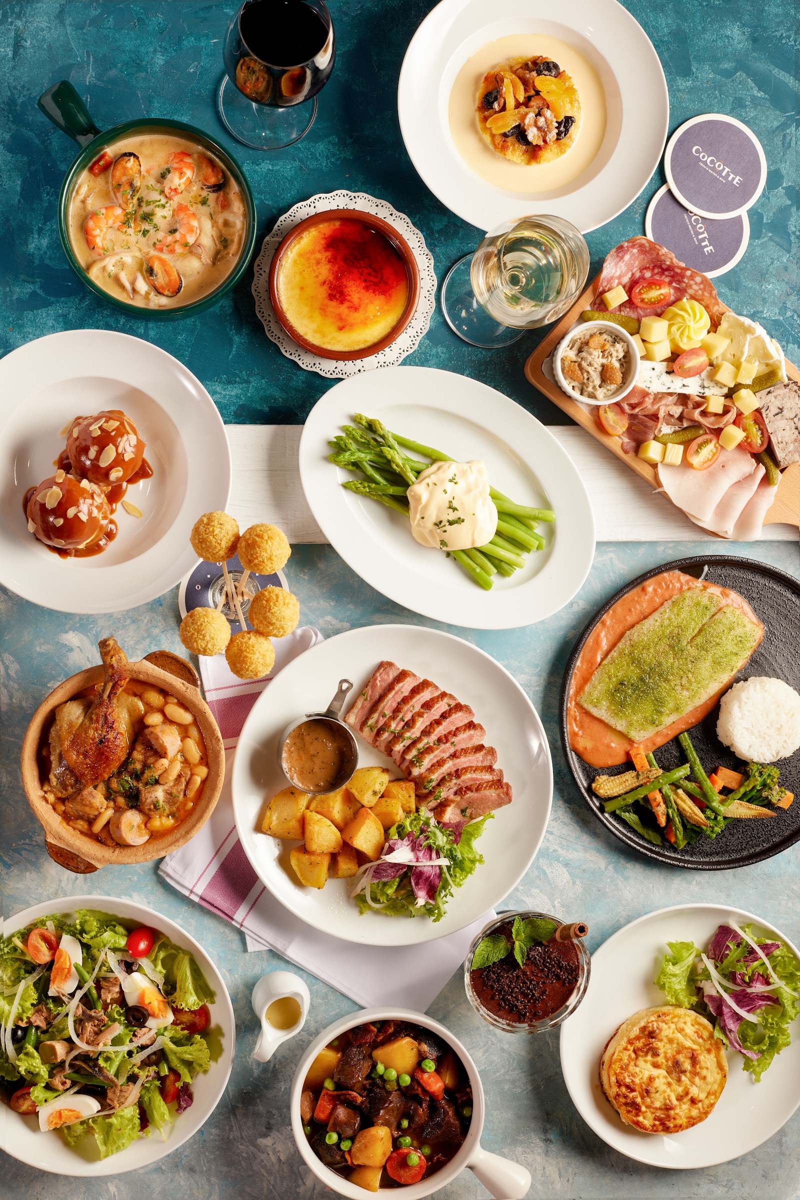 Bàn tiệc nhìn từ trên xuống: nồi cocotte hải sản, nồi bò hầm, khay phô mai và thịt nguội, măng tây, cá hồi, vịt thái lát, salad, bánh và rượu vang