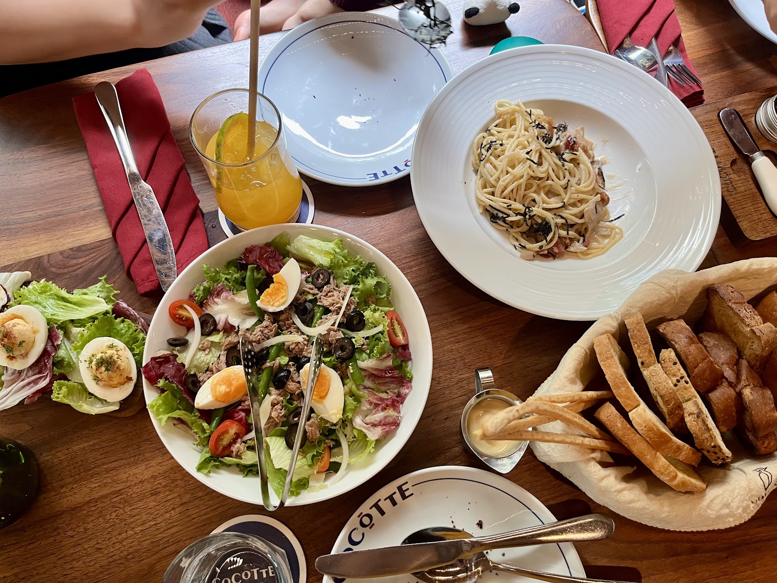 Bàn tiệc nhìn từ trên xuống: salade niçoise với trứng và cá ngừ, đĩa pasta, giỏ bánh mì, đĩa in tên CoCOTTE và khăn ăn đỏ