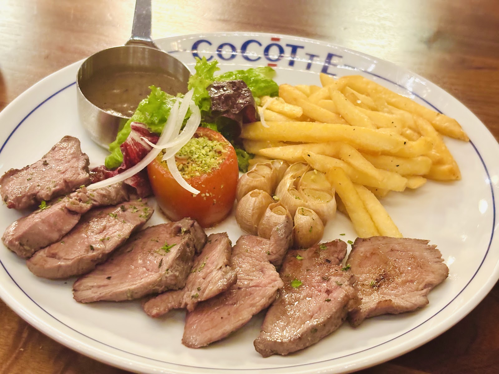 Steak thái lát với khoai chiên vàng, tỏi nướng, salad và chén sốt tiêu trên đĩa in tên CoCOTTE
