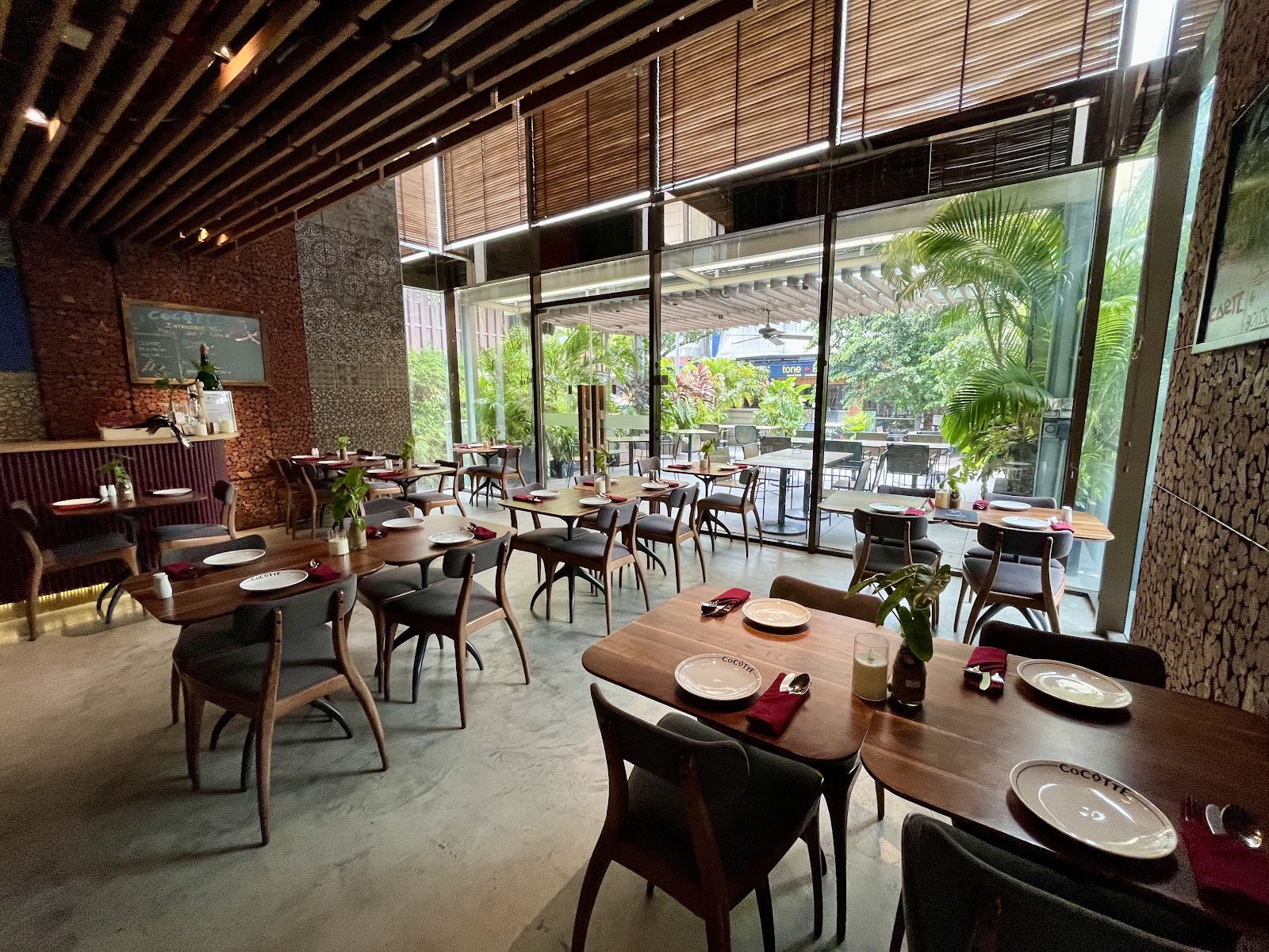 Phòng ăn trần gỗ với bàn bistro khăn đỏ và đĩa CoCOTTE; cả bức tường kính mở ra terrace sân vườn xanh bên ngoài