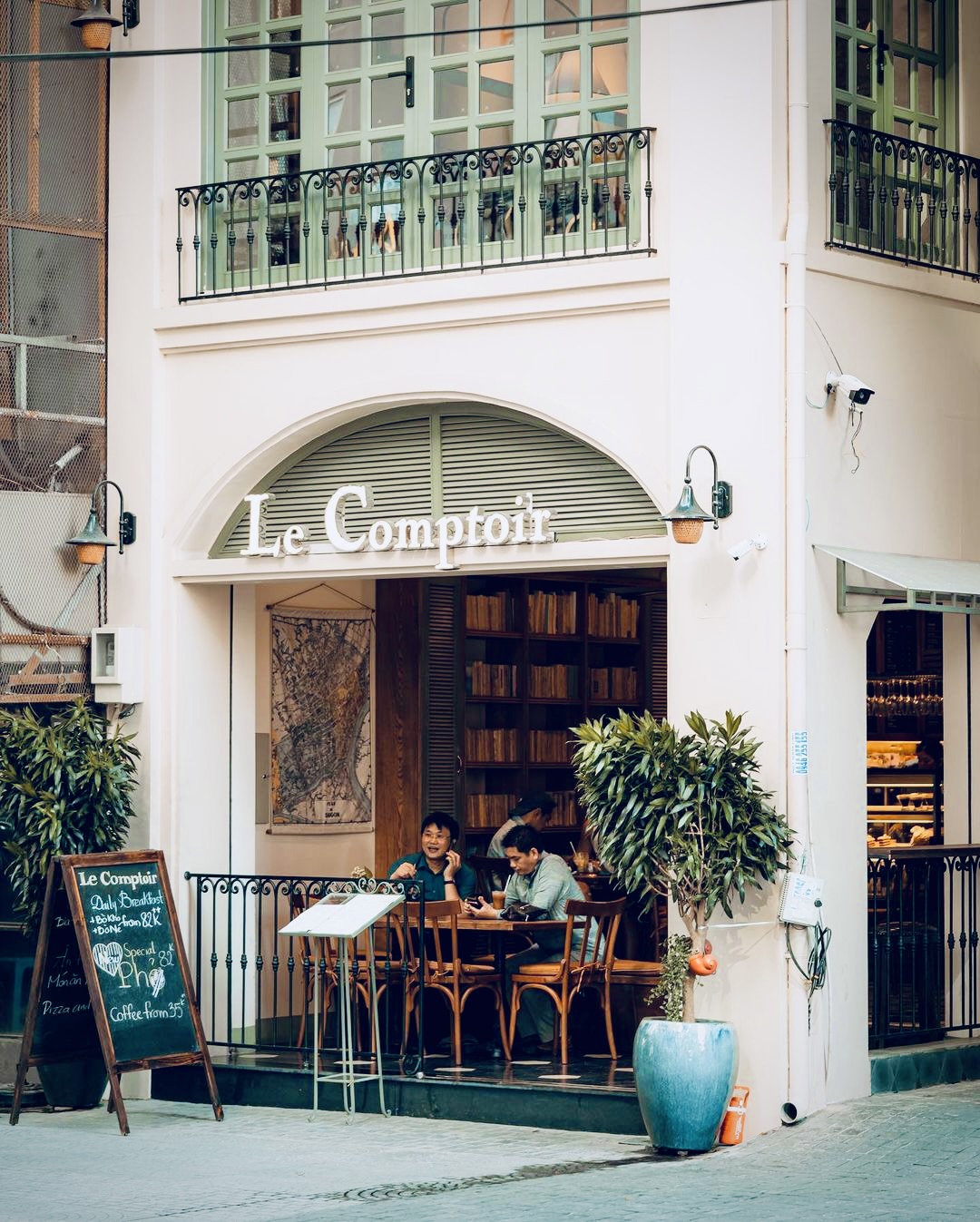 Mặt tiền Le Comptoir ở 12 Hồ Huấn Nghiệp — biển chữ serif trắng trên cửa vòm, cửa chớp xanh xô thơm, bàn ngoài trời có khách
