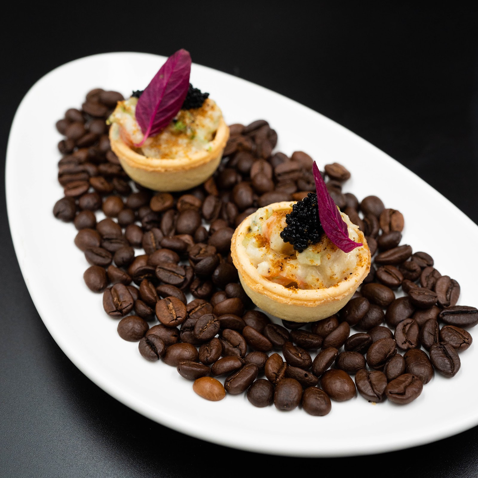 Hai chiếc bánh tart hải sản phủ trứng cá đen và lá tía tô đỏ, dọn trên đĩa trắng phủ hạt cà phê