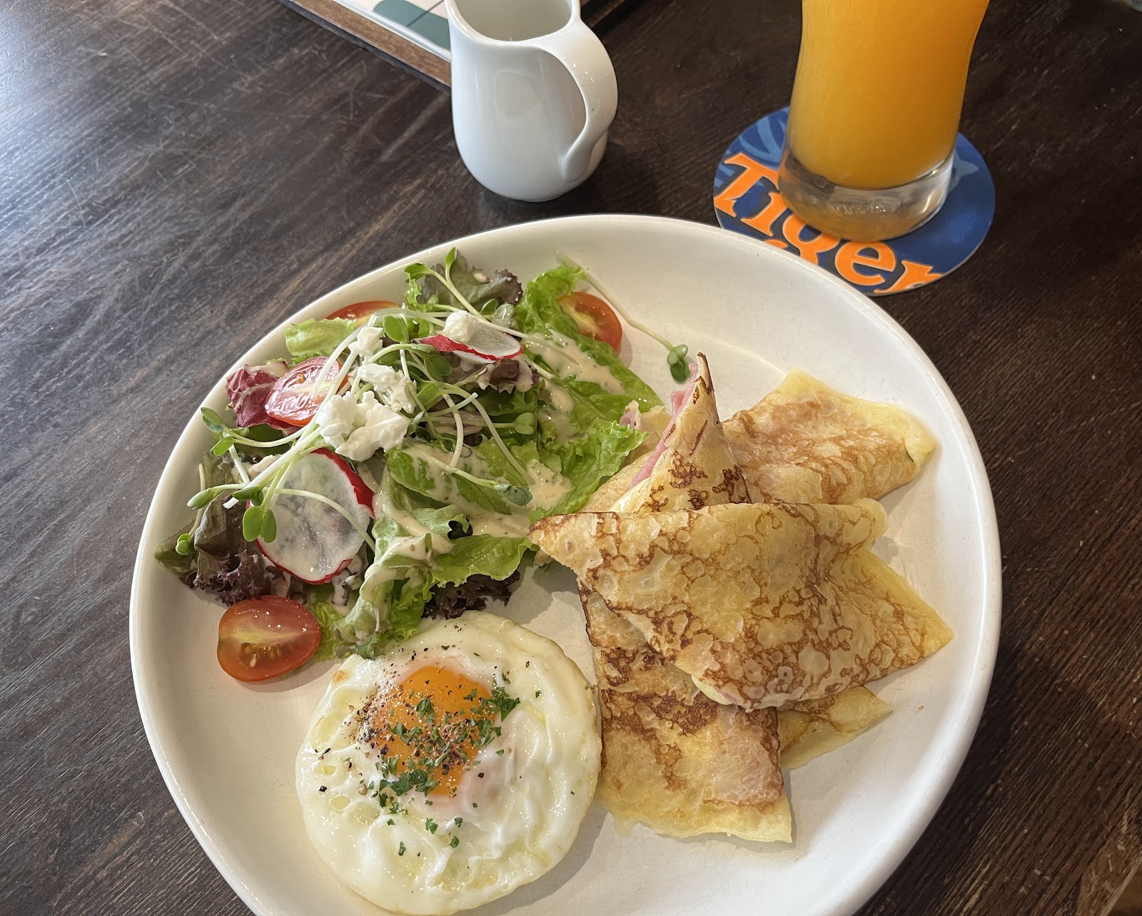 Đĩa crêpe jambon & phô mai gấp nếp kèm trứng ốp la, salad xanh trộn với củ cải và cà chua, ly nước cam trên bàn gỗ sẫm