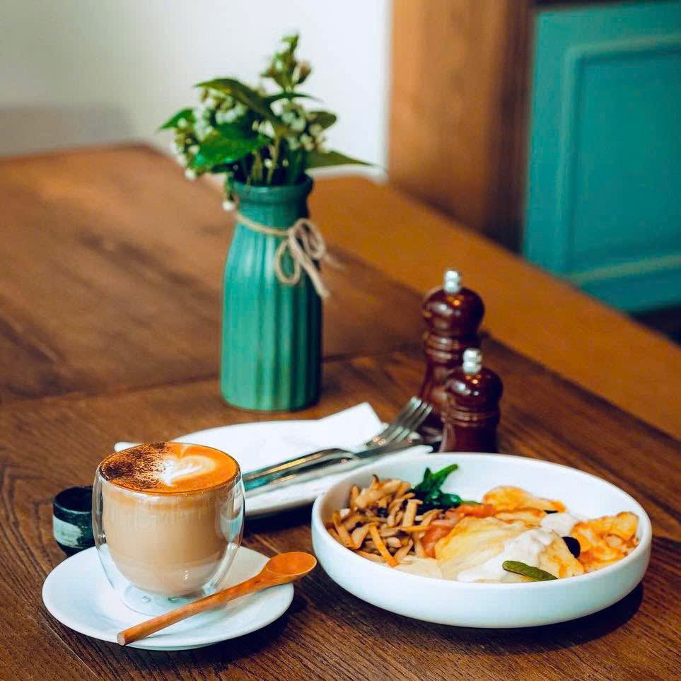 Ly latte thủy tinh rắc cacao cạnh tô điểm tâm trắng đựng cá hồi, trứng và nấm trên bàn gỗ, lọ hoa phía sau