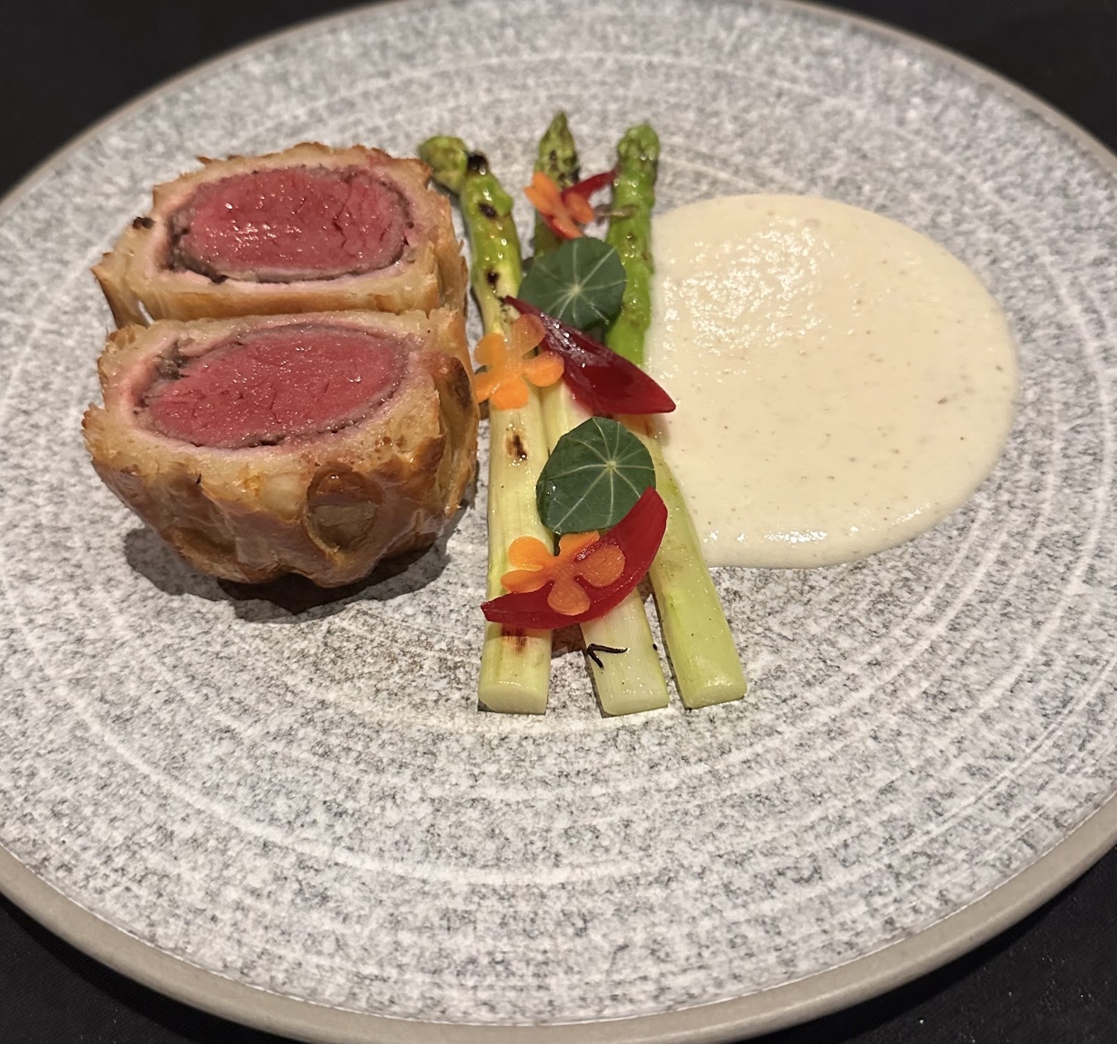 Beef wellington cross-section — golden pastry around the pink fillet — with charred asparagus, a white cream smear and red wine on the grey speckled plate