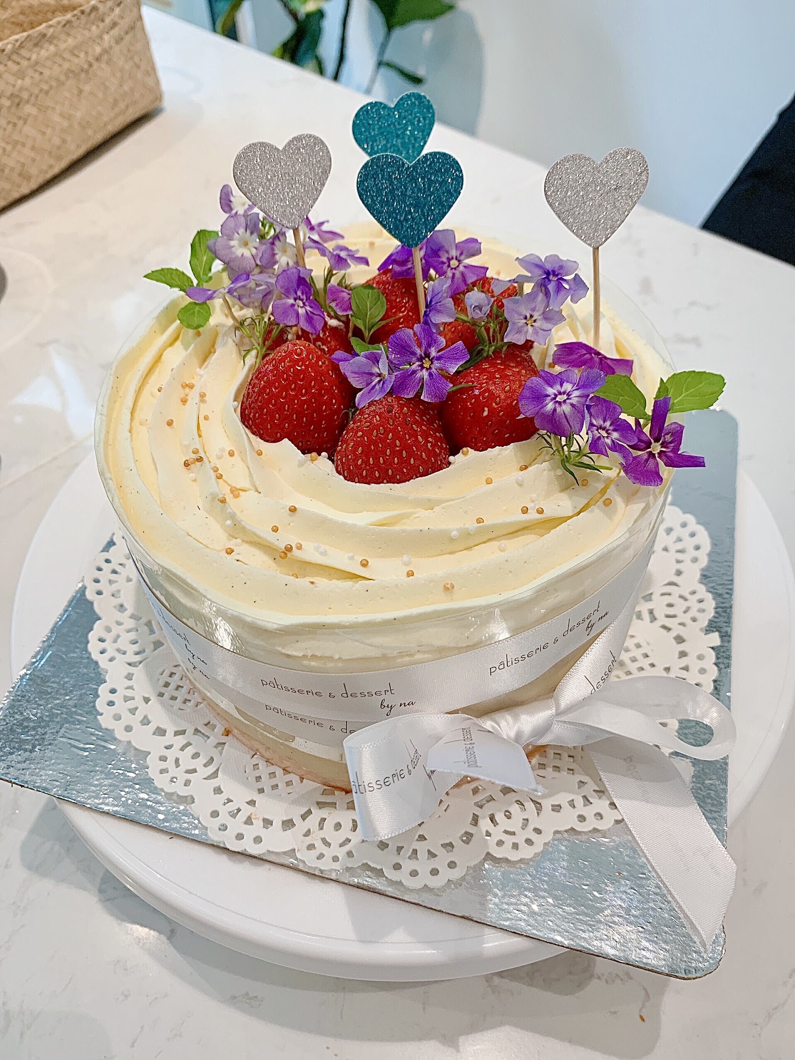 Strawberry celebration cake with violet flowers and heart picks — the ribbon reads 'patisserie & dessert by na'