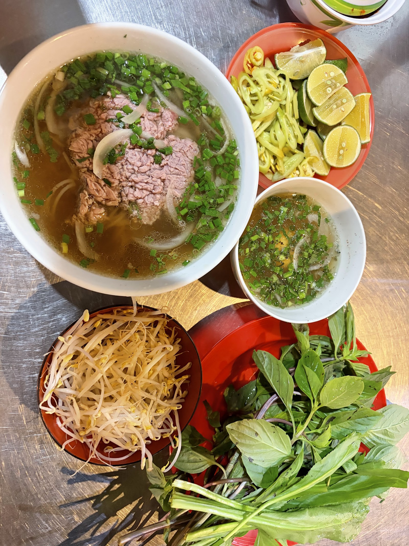 Bàn ca tối: tô tái đầy, chén giá, chanh ớt, đĩa rau thơm — dọn trên mặt inox