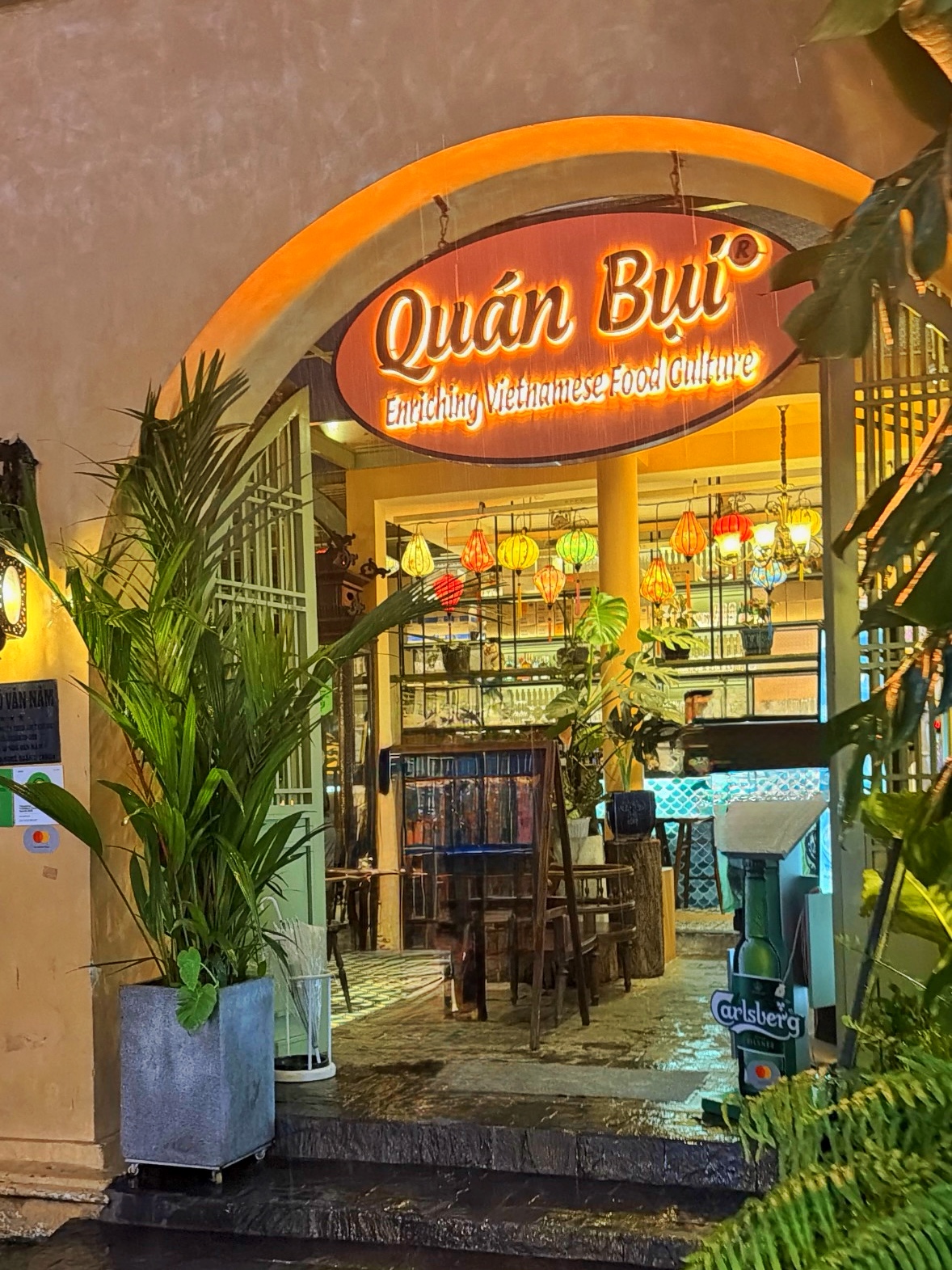 Cửa vào Quán Bụi — cây xanh và đèn ấm chào đón thực khách