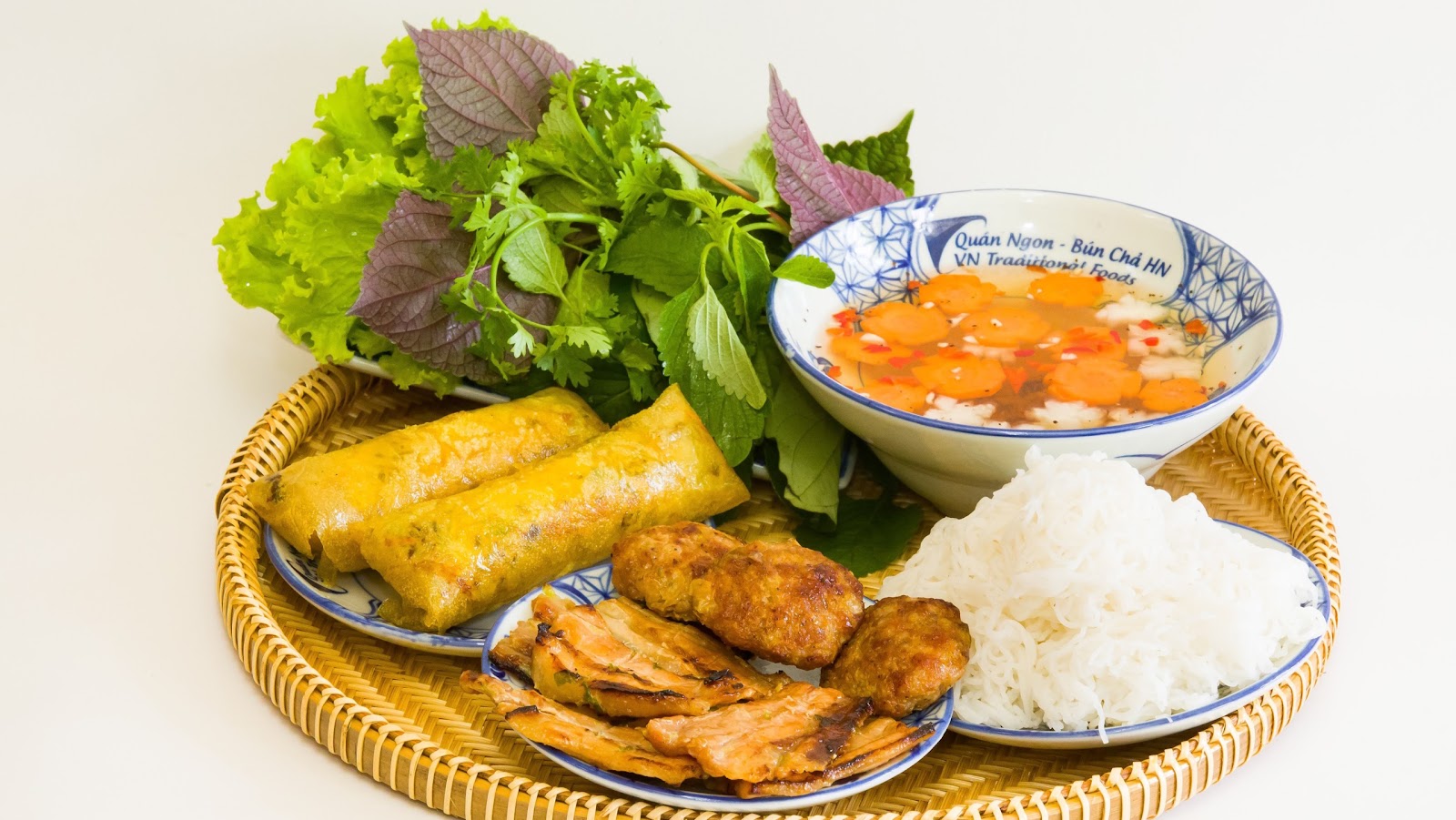 The owner's bún chả platter: the house-branded dipping bowl, carrot flowers, grilled pork patties and belly, fried rolls and vermicelli on a round rattan tray