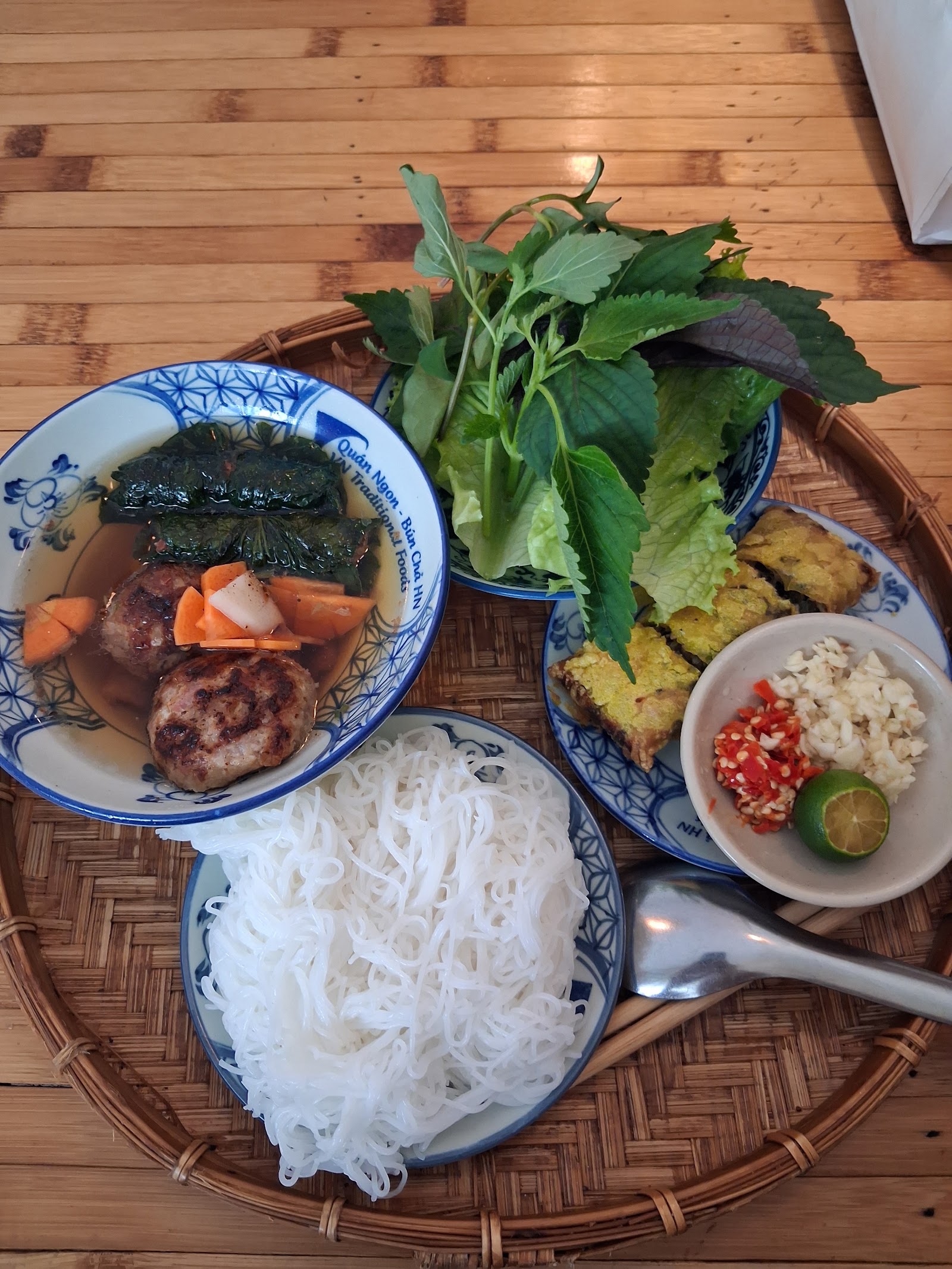 A bún chả set with dipping sauce, pork patties, fried rolls, vermicelli and herbs on a rattan tray