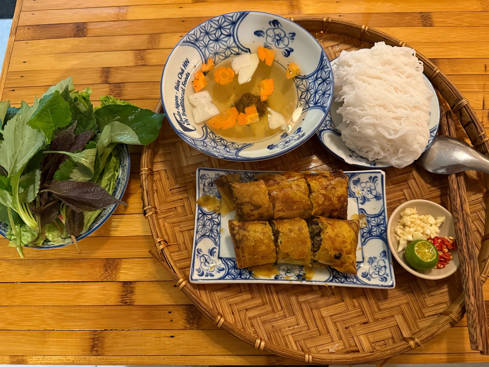 A bún chả platter with crispy fried rolls, a carrot-flower dipping bowl and fresh herbs
