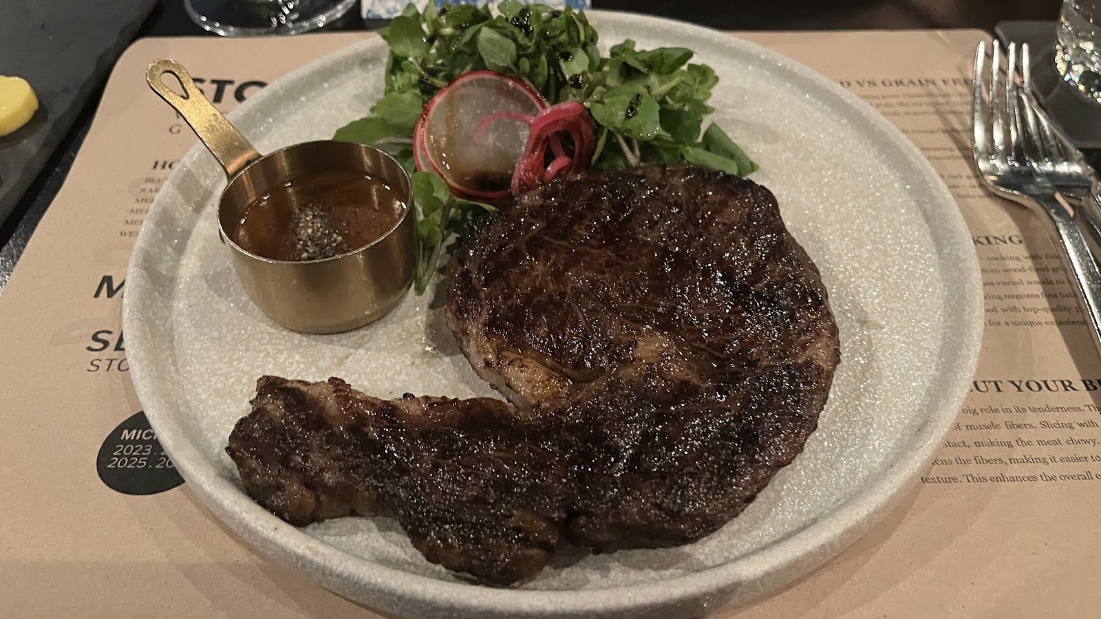 Miếng steak nướng vỉ trên đĩa đá, kèm hũ sốt đồng và rau xanh
