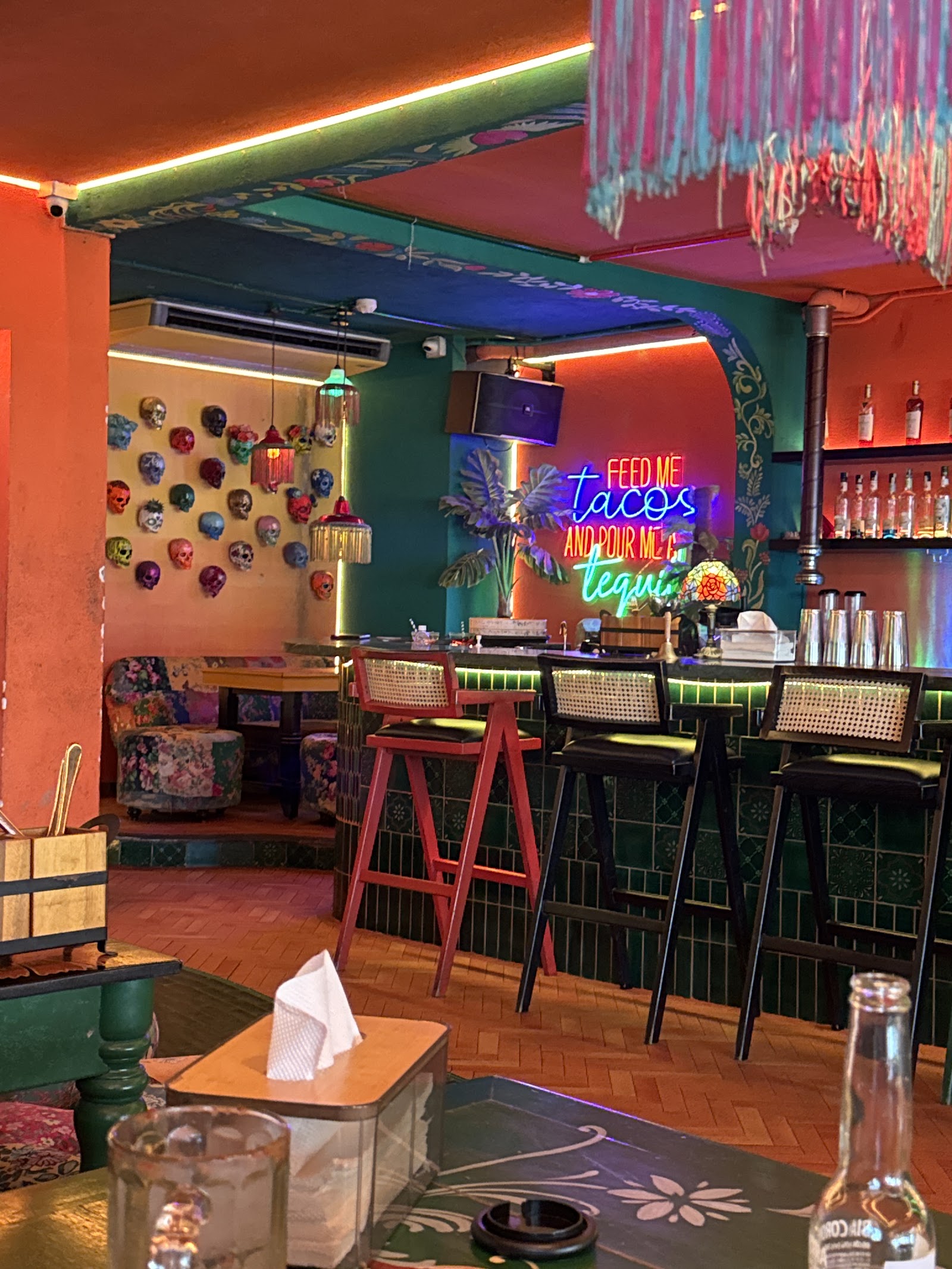 Quầy bar với biển neon “FEED ME tacos” và tường mặt nạ calavera