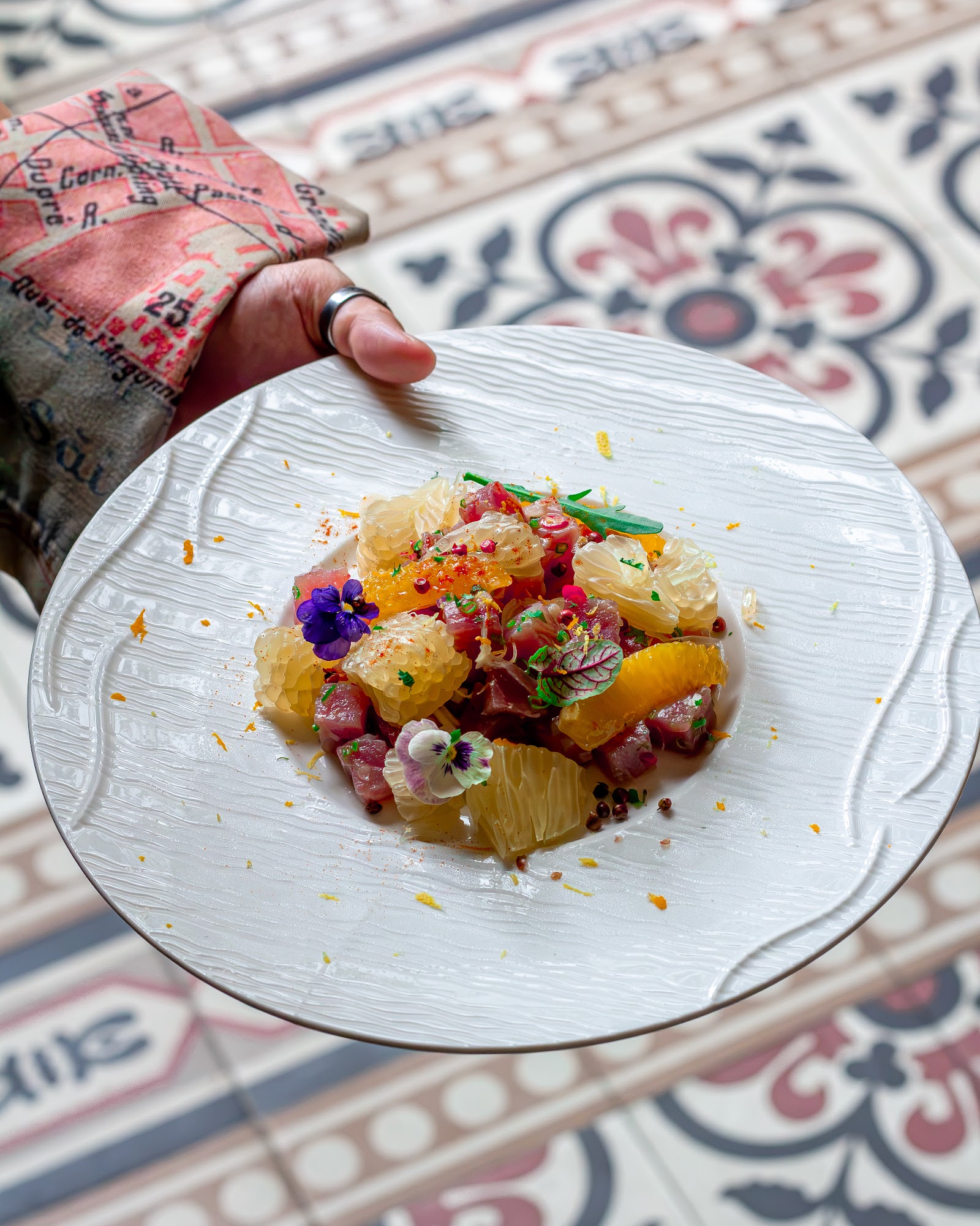 Tartare cá với múi bưởi và cam, hoa ăn được tím và trắng, lá thơm — đĩa trắng đặt trên nền gạch bông hoa văn